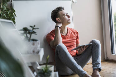 Nachdenklicher junger Mann mit digitalem Tablet, der wegschaut, während er im Wohnzimmer sitzt - UUF22777