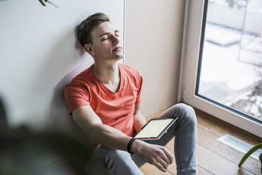 Junger Mann mit digitalem Tablet, der sich entspannt, während er im Wohnzimmer an der Wand sitzt - UUF22778