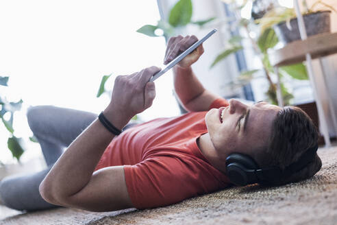 Mann sieht sich ein Video auf einem digitalen Tablet an, während er auf dem Teppich im Wohnzimmer liegt - UUF22782