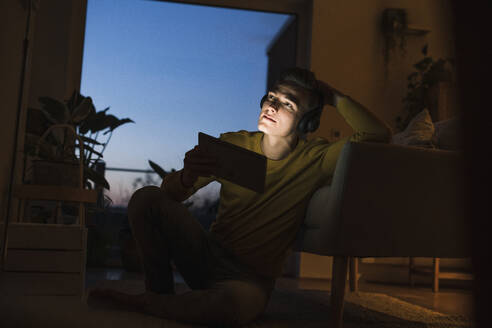 Nachdenklicher junger Mann mit Kopfhörern und digitalem Tablet, der im Wohnzimmer wegschaut - UUF22795
