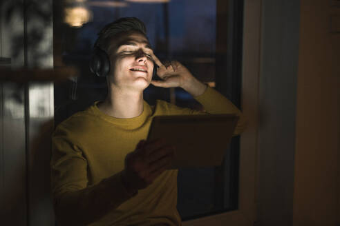 Young man with digital tablet listening music through wireless headphones while sitting in living room - UUF22796