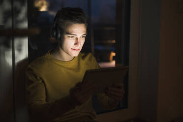 Junger Mann trägt Kopfhörer, während er im Wohnzimmer Videos auf einem digitalen Tablet anschaut - UUF22797