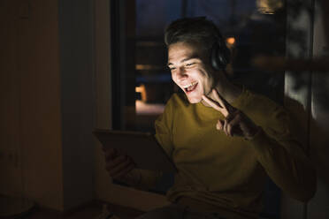 Cheerful man wearing headphones gesturing peace sign on video call while sitting in living room - UUF22798