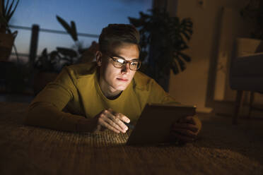 Junger Mann mit Brille benutzt ein digitales Tablet, während er nachts auf dem Teppich im Wohnzimmer liegt - UUF22804