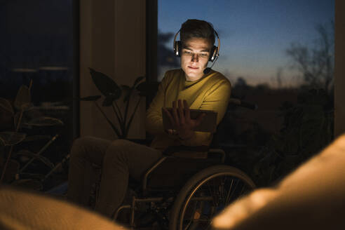 Junger Mann im Rollstuhl, der drahtlose Kopfhörer trägt, während er im Wohnzimmer auf dem Rollstuhl sitzend ein digitales Tablet benutzt - UUF22808
