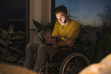 Behinderter junger Mann, der im Rollstuhl sitzend im Wohnzimmer ein digitales Tablet benutzt - UUF22811