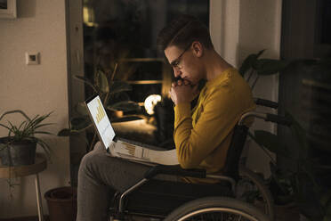 Concentrated oung man in wheelchair with laptop working late while sitting on wheelchair in living room - UUF22812