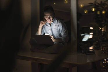 Lächelnder Geschäftsmann mit Laptop, der ein Smartphone benutzt, während er auf einem Tisch im Wohnzimmer sitzt - UUF22819