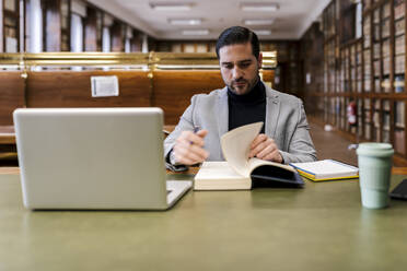 Mittlerer Erwachsener Geschäftsmann mit Laptop, der in einer Bibliothek sitzt und ein Buch liest - EGAF01898