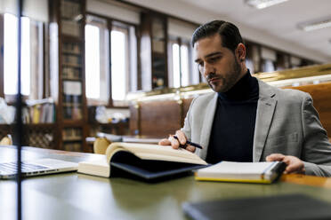 Geschäftsmann mit Laptop, der ein Buch liest, während er in einer Bibliothek arbeitet - EGAF01899