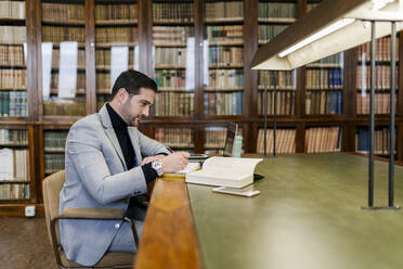 Geschäftsmann mit Laptop, der in einem Buch schreibt, während er in einer Bibliothek sitzt - EGAF01902