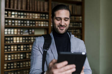 Geschäftsmann lächelnd mit digitalem Tablet in einer Bibliothek stehend - EGAF01907