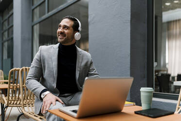 Lächelnder Geschäftsmann mit Laptop und Kopfhörern, der wegschaut, während er im Café sitzt - EGAF01908