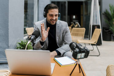 Smiling businessman waving hand toward camera while vlogging at cafe - EGAF01923