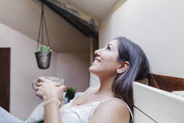 Smiling woman with coffee cup looking up while leaning on bed at home - EBBF02541