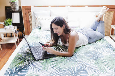 Smiling woman with laptop using mobile phone while lying on bed at home - EBBF02546