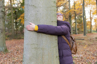 Lächelnde Frau schaut nach oben, während sie einen Baum im Wald umarmt - WPEF04192