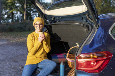 Lächelnde Frau, die ein heißes Getränk zu sich nimmt, während sie im Kofferraum eines Autos sitzt - WPEF04198