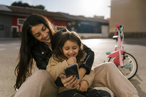 Smiling mother using mobile phone while sitting on road - EGAF01928