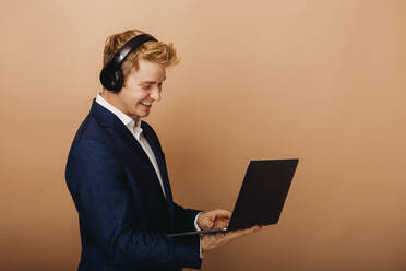 Lächelnder Geschäftsmann mit drahtlosen Kopfhörern und Laptop vor braunem Hintergrund - DAWF01863
