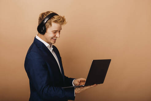 Smiling businessman with wireless headphones using laptop by brown background - DAWF01863