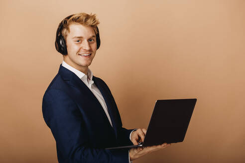 Smiling male entrepreneur with headphones holding laptop by brown background - DAWF01864
