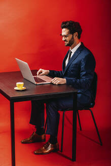 Lächelnder Geschäftsmann mit Laptop am Schreibtisch vor rotem Hintergrund - DAWF01874