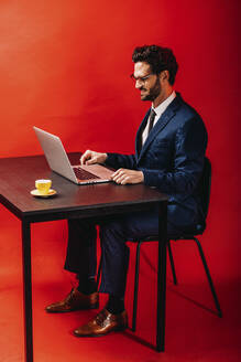 Smiling businessman using laptop at desk by red background - DAWF01874