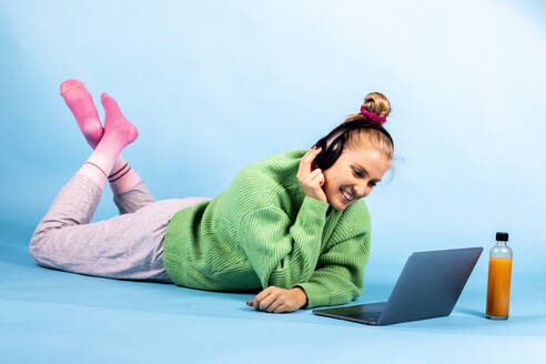 Lächelnde Frau mit Kopfhörern, die auf einen Laptop schaut, während sie auf der Vorderseite vor blauem Hintergrund liegt - DAWF01876