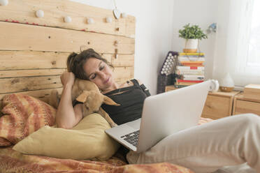 Frau mit Laptop, die ihren Hund umarmt, während sie zu Hause auf dem Bett sitzt - SKCF00727