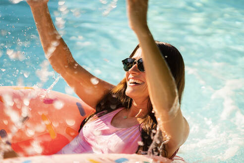 Cheerful woman with inflatable ring splashing water while enjoying in swimming pool - AKLF00079