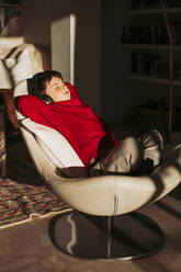 Junge hört Musik über Kopfhörer, während er sich auf einem Stuhl zu Hause entspannt - LJF02067