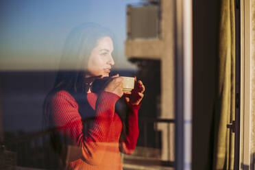 Nachdenkliche Frau, die einen Kaffee trinkt, während sie durch ein Fenster zu Hause schaut - LJF02076