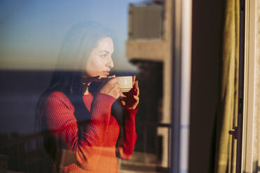 Frau trinkt Kaffee und schaut durch das Fenster zu Hause - LJF02077