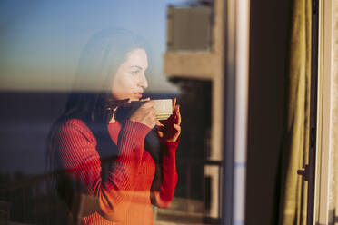 Woman having coffee while looking through window at home - LJF02077