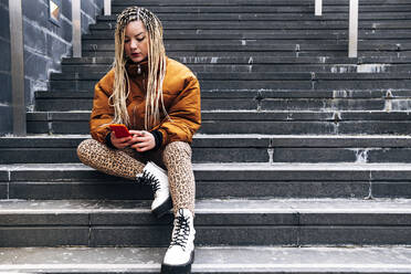 Junge Frau in Jacke, die auf einer Treppe sitzend ein Mobiltelefon benutzt - ASGF00027