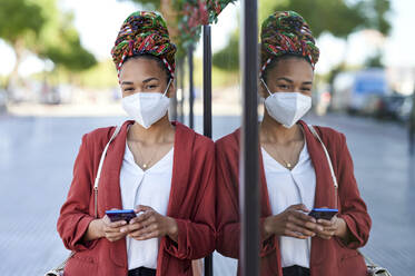 Frau mit Kopftuch und Gesichtsmaske benutzt ein Mobiltelefon, während sie sich an eine Glaswand lehnt - KIJF03616