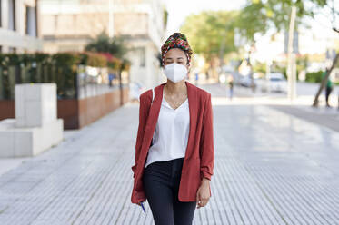 Young woman wearing headscarf and protective face mask walking on footpath - KIJF03619