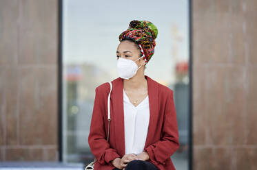 Woman with headscarf and protective face mask looking away while sitting outdoors - KIJF03621