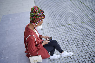 Woman wearing protective face mask and headscarf using mobile phone while sitting on footpath - KIJF03623