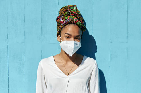 Woman wearing protective face mask and headscarf staring while standing against blue wall - KIJF03633