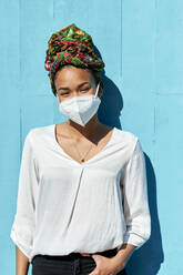 Junge Frau mit Kopftuch und Gesichtsmaske steht mit den Händen in den Taschen vor einer blauen Wand - KIJF03634