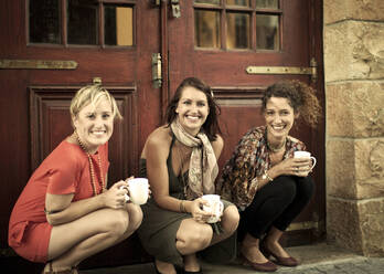 Young women with coffee cup smiling while crouching against door - AJOF01128