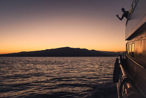A man's silhouette shows shaka on sunset cruise with Maui in distance - CAVF93488