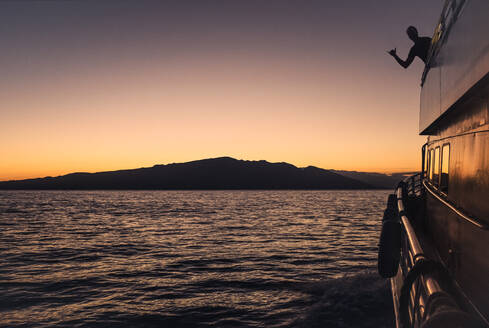 Die Silhouette eines Mannes zeigt einen Shaka auf einer Kreuzfahrt bei Sonnenuntergang mit Maui in der Ferne - CAVF93488