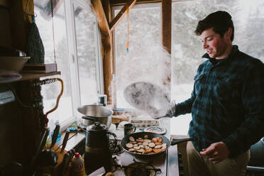 A young man cooks steaming breakfast in a cabin during winter - CAVF93529