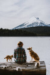 A woman sits and enjoys view of lake and mountains with her two dogs - CAVF93534