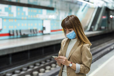 Woman with protective face mask using mobile phone while standing at subway station during pandemic - AMPF00043