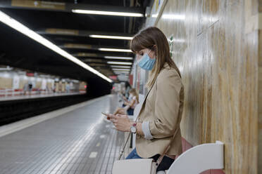 Woman with using mobile phone while sitting at subway platform during COVID-19 - AMPF00046