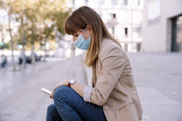 Young woman with protective face mask using smart phone while sitting outside - AMPF00048